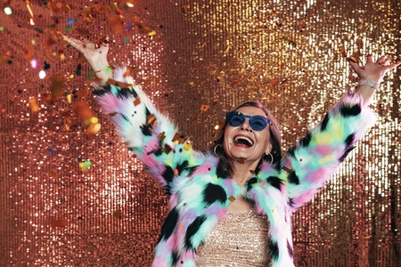 Happy senior woman raising hands up while having fun under confetti against glitter background