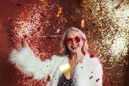 Happy senior woman in pink eyeglasses and white fur coat having fun under confetti