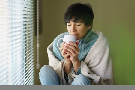 Middle aged woman smelling coffee from her cup near a window