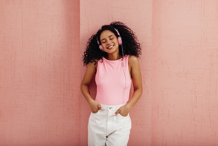 Smiling woman leaning on pink wall listening to music with closed eyes