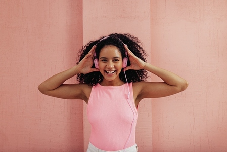Cheerful woman on pink headphones looking at camera standing against pink wall