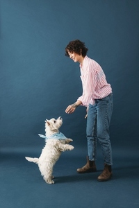 Side view of young woman in casuals standing in studio on blue background and feeding her dog