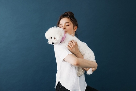 Young woman holding her little white dog in studio over blue background