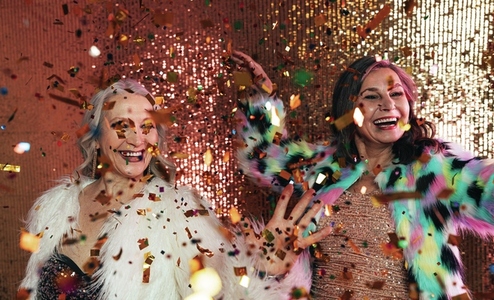 Senior females in fur coats celebrating together in studio under colorful confetti