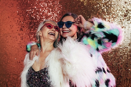 Two women wearing eyeglasses and fur coats  Senior females laughing against glitter background