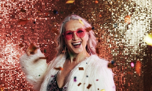 Laughing woman in white fur coat throwing confetti in studio against glitter background