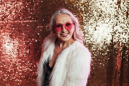 Stylish senior woman in white fur coat wearing pink eyeglasses having fun in studio