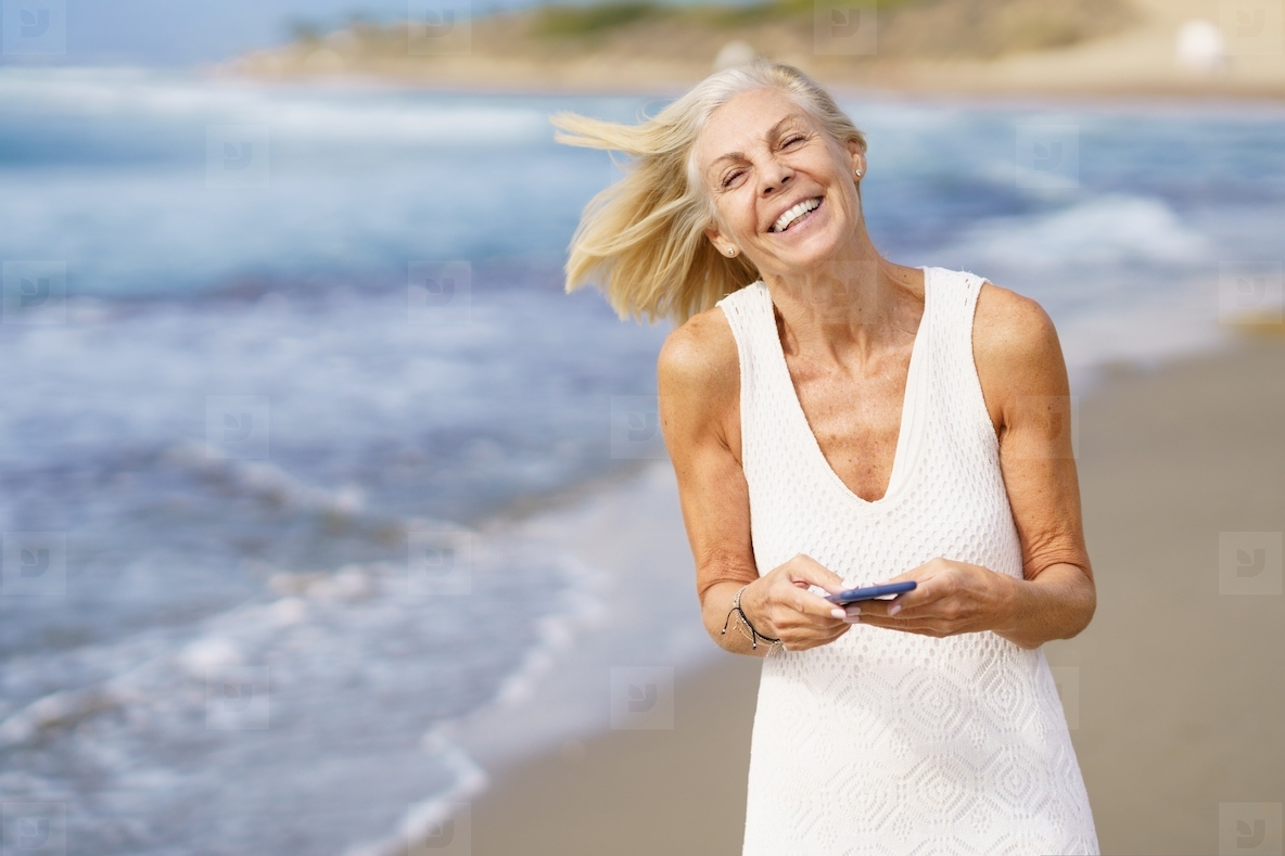 Smiling mature woman walking on the beach using a smartphone