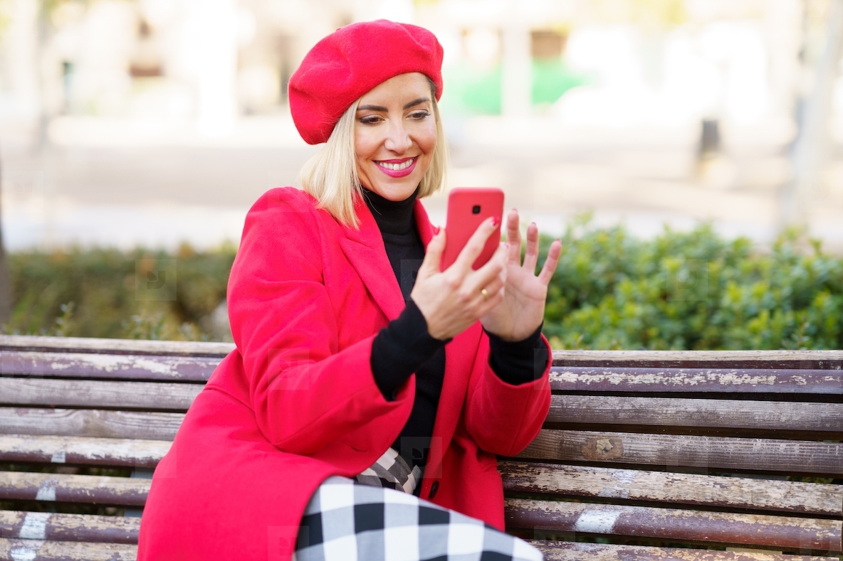 Middle aged woman sitting on park bench using smartphone wearing red winter clothes
