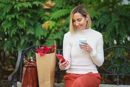Woman sitting on park bench using smartphone while drinking hot coffee in a take away glass
