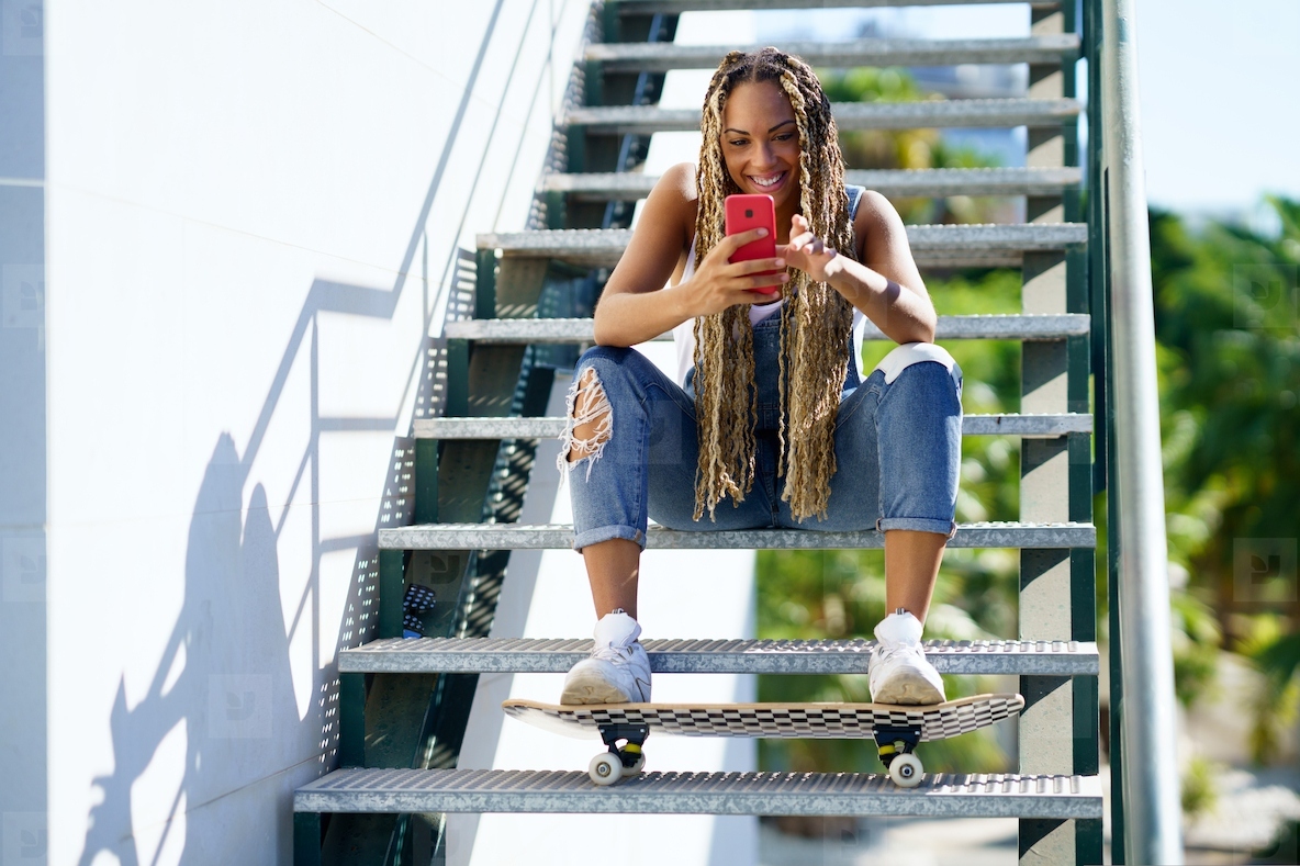 Black woman with coloured braids  consulting her smartphone with her feet resting on a skateboard