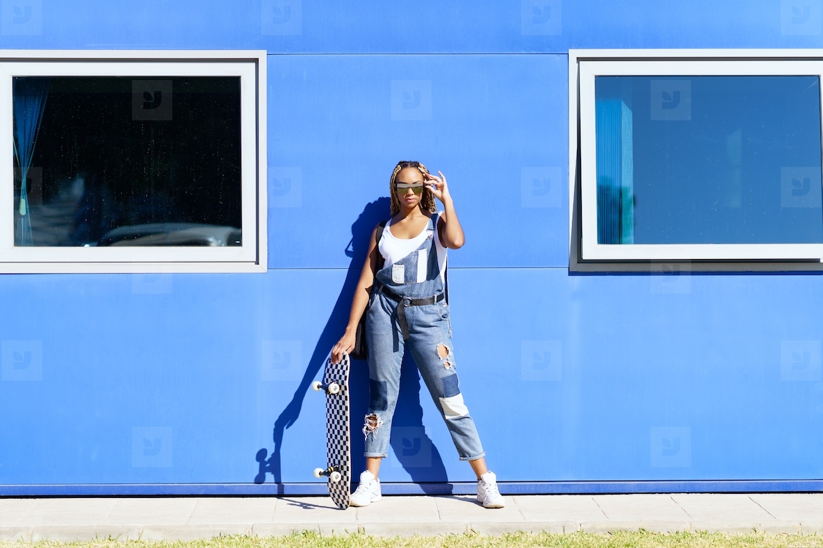 Black girl dressed casual  with modern sunglasses and a skateboard on blue wall background