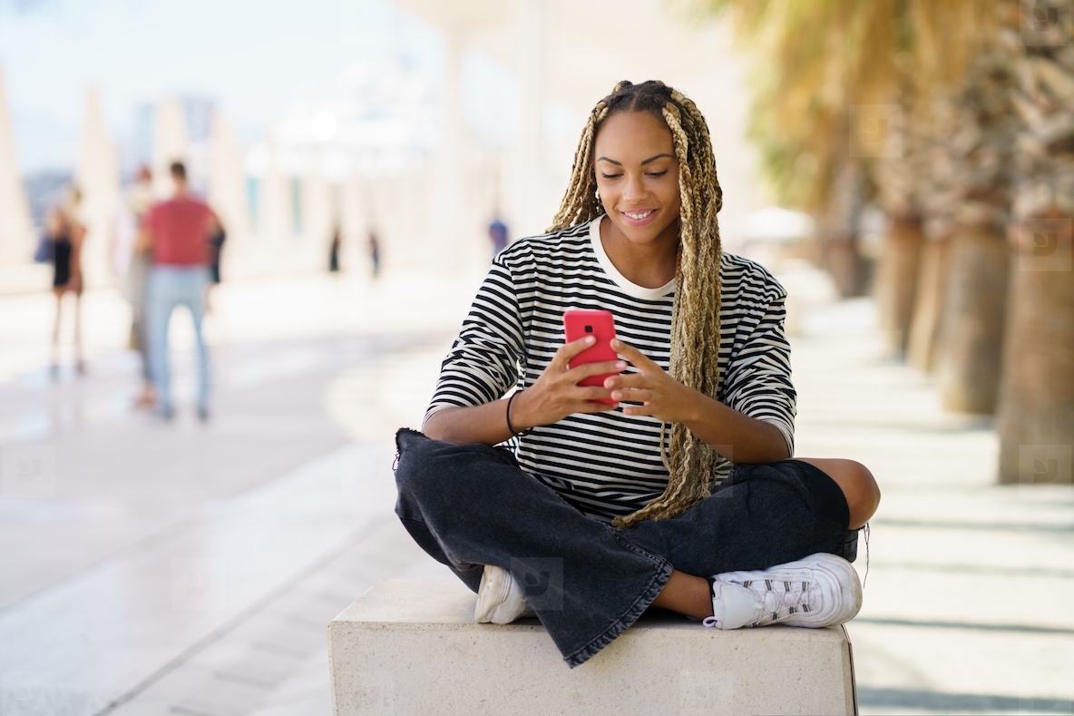 Black woman texting with a smartphone sitting on a bench outdoors  wearing her hair in braids