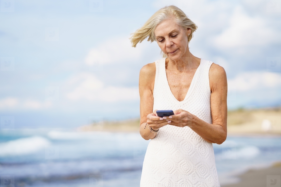Senior woman walking on the beach using a smartphone