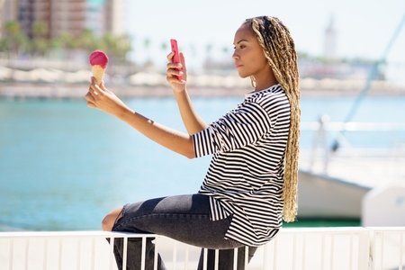 Black girl photographing with her smartphone a strawberry ice cream she is eating near the sea