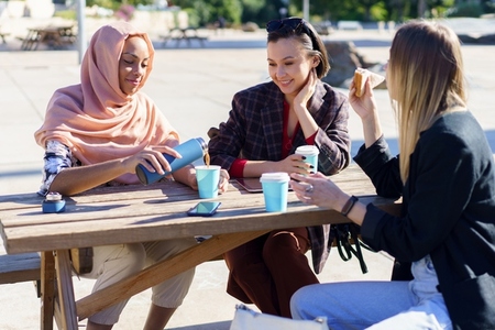 Positive diverse women having coffee break at table in park