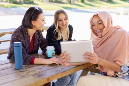 Cheerful diverse ladies sharing tablet during coffee break in cafe