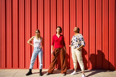 Stylish young diverse ladies standing against red fence on street