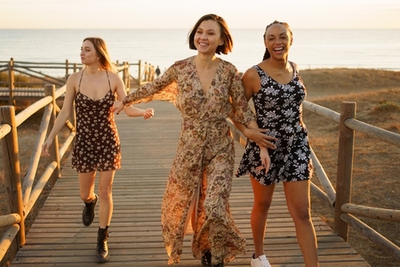 Smiling multiethnic girlfriends walking on wooden boardwalk on sea shore