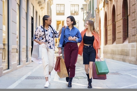 Stylish multiracial female shopaholics walking together on street with paper bags