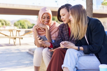 Positive multiethnic girlfriends sharing smartphone while resting on bench in park