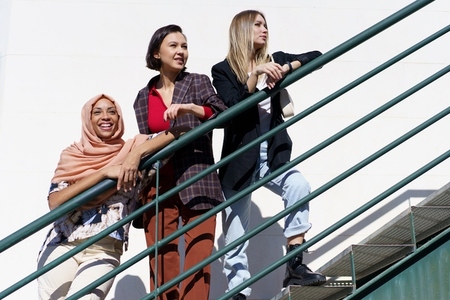 Content young diverse ladies looking away while standing on stairs in city