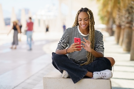 Black girl texting with a smartphone sitting on a bench outdoors  wearing her hair in braids
