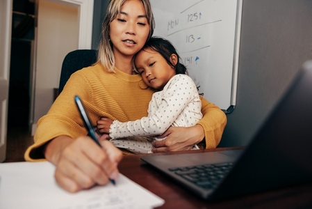 Young working mom writing notes while embracing her daughter