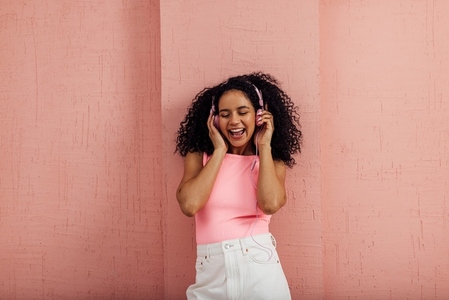 Happy woman with curly hair holding pink headphones with closed eyes leaning a wall