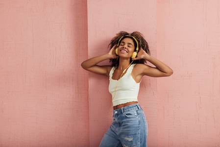 Cheerful woman wearing casual clothes and yellow headphones standing at pink wall