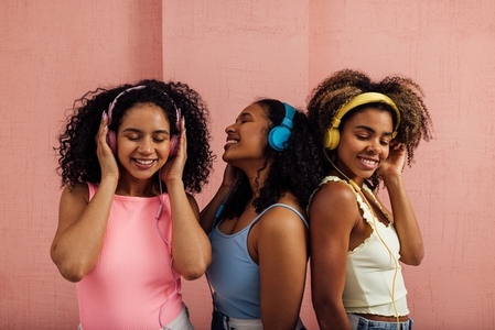 Group of female friends in casuals wearing headphones standing together at pink wall