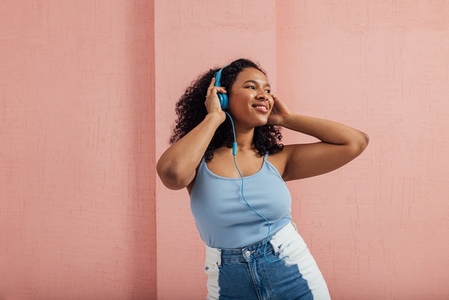 Smiling woman in casuals standing against pink wall listening to music and looking away
