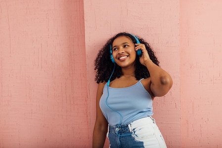 Happy woman wearing blue headphones leaning a pink wall