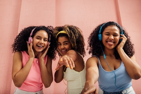Three laughing women with curly hair dancing together and looking at camera  wearing headphones