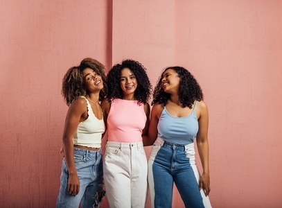 Female friends with curly hair posing together against pink wall