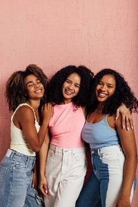 Three smiling women with curly hair standing together and looking at camera against a pink wall
