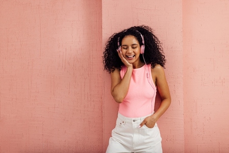 Cheerful woman listening to music leaning pink wall with closed eyes