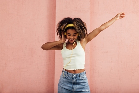 Cheerful woman with curly hair wearing yellow headphones dancing against a wall