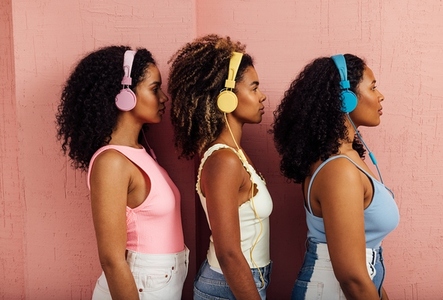 Side view of three women wearing headphones standing one after another against a pink wall