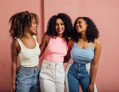 Three cheerful women with curly hair having fun together while standing at a pink wall