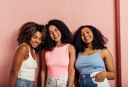 Portrait of a three smiling women with curly hair posing together against pink wall looking at camera