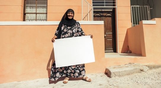 Cheerful Muslim woman holding a blank placard outside her home