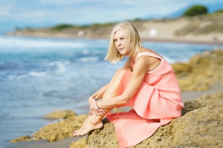 Mature woman sitting on some rocks on the shore of a tropical beach  wearing a nice orange dress