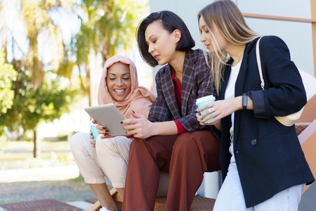 Stylish diverse women friends sharing tablet and drinking takeaway coffee on street