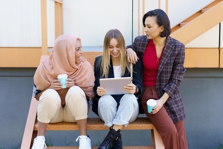 Stylish diverse female friends sharing tablet and drinking takeaway coffee on street