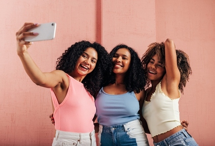 Three happy women with curly hair taking selfie against pink wall
