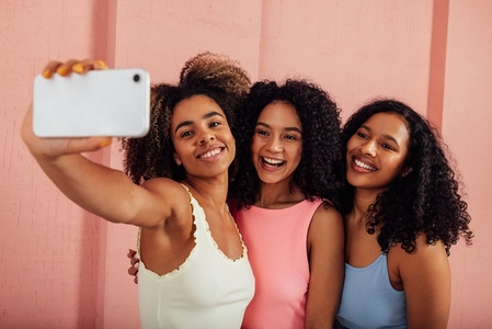 Young women in bright casuals standing together and taking selfie on a mobile phone  Happy females having fun against pink wall outdoors