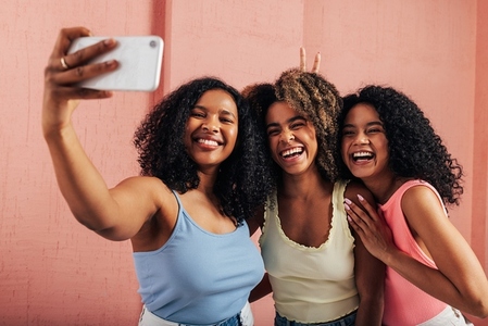 Three women with curly hair laughing together while taking selfie against a pink wall