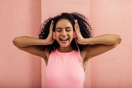 Laughing woman wearing pink headphones enjoying music with closed eyes  Smiling female with curly hair against a pink wall listening to music