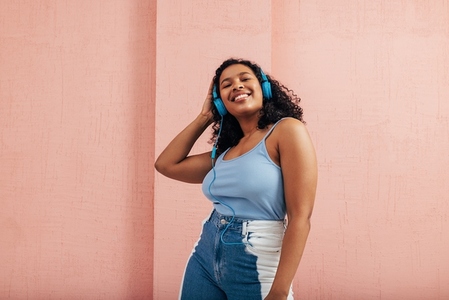 Cheerful woman with black curly hair wearing blue headphones standing at pink wall